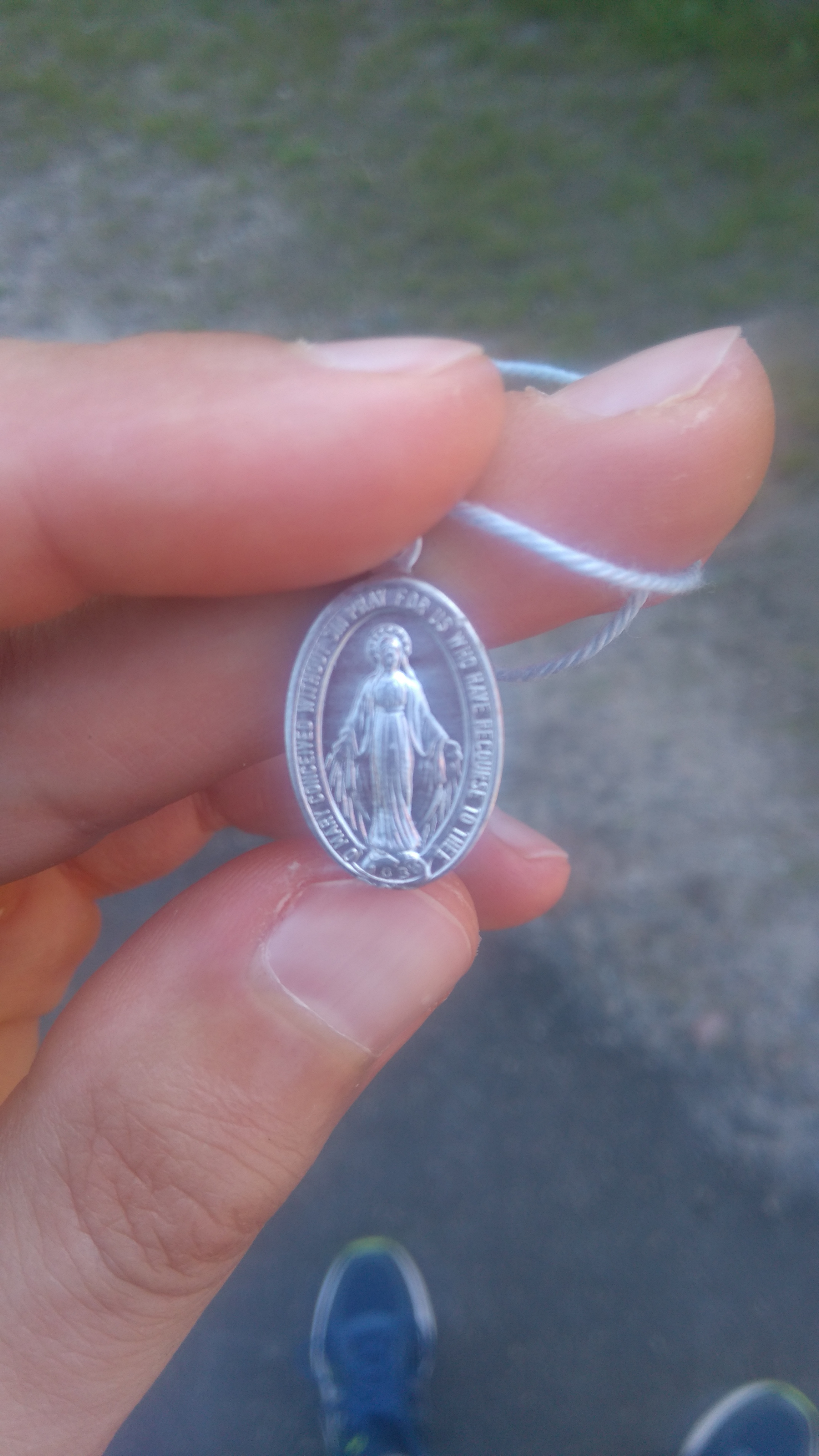 Hopeanvärinen ihmeitätekevä medaljonki vasemman käden etusormen ja peukalon välissä, etupuoli (Neitsyt Maria)