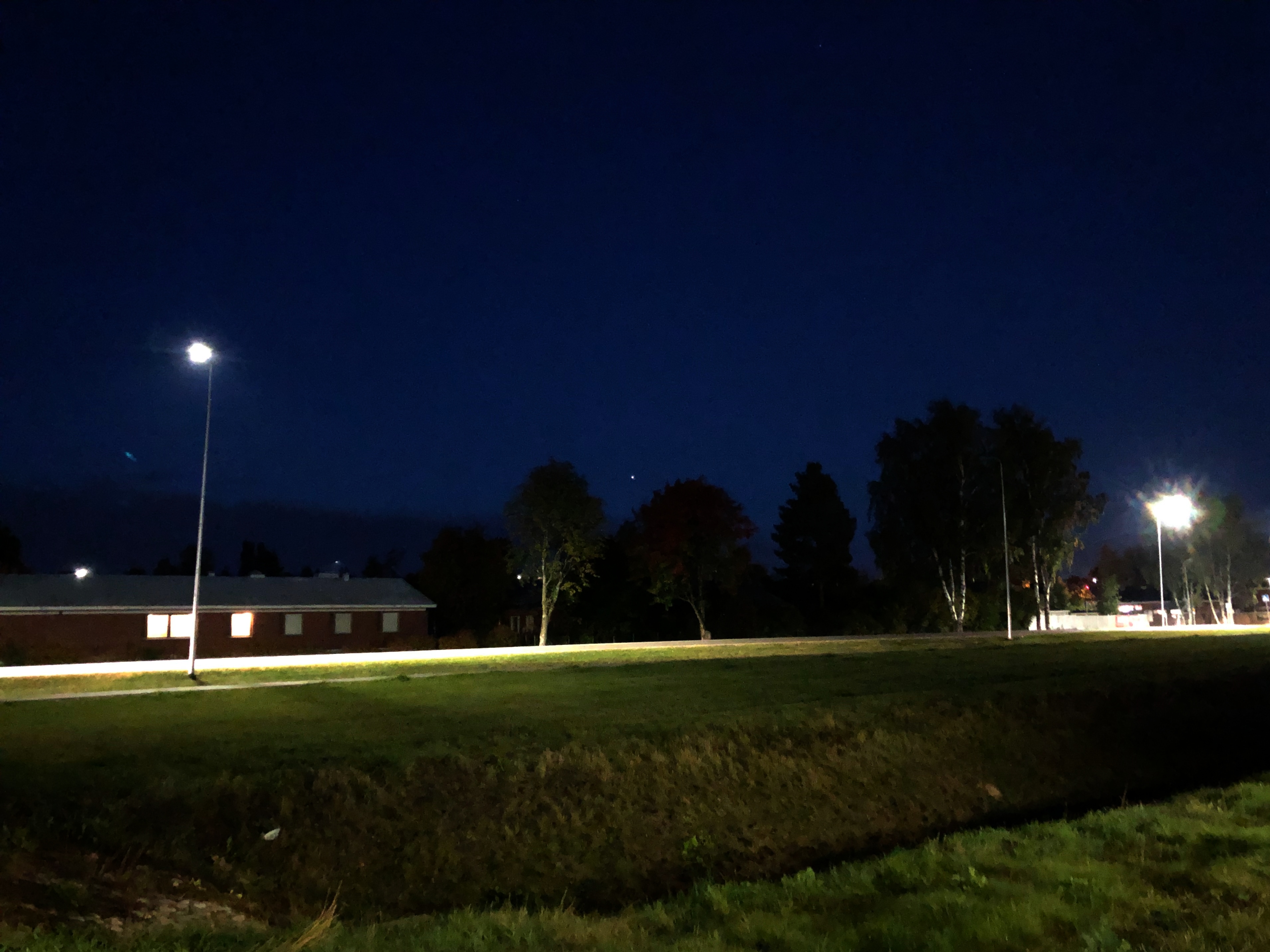 Tummansininen iltahämärä. Taivaanrannassa (keskellä kuvaa) valopiste (Jupiter). Katuvalojen valaisema tie, ja etualalla vihreää ruohoa sekä oja.