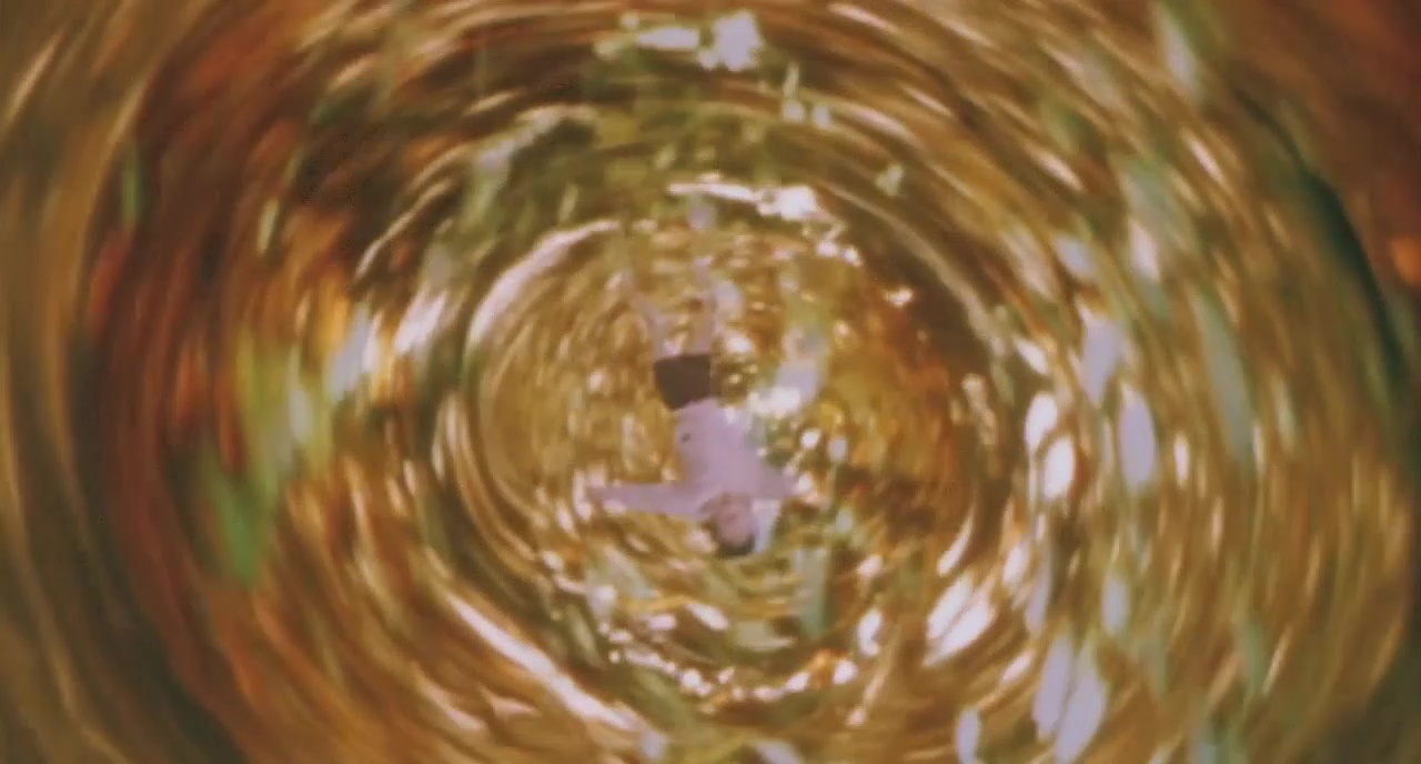 Kullankirjava, nestepyörteen näköinen tunneli. Keskellä ylösalaisin lapsi, jolla valkoinen paita ja tummat sortsit.