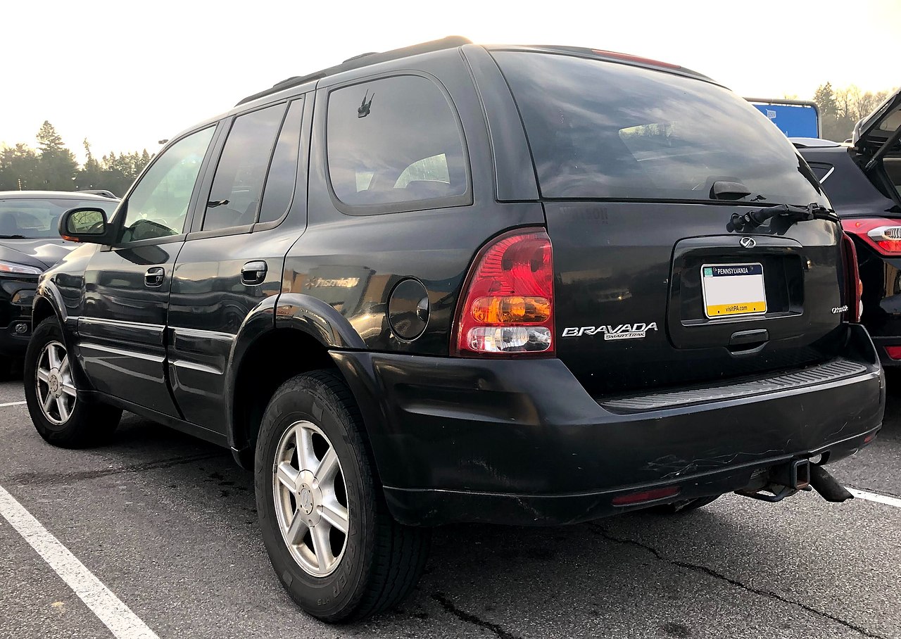 Pysäköintiruutuun pysäköity musta katumaasturi (Oldsmobile Bravada vm. 2002, Pennsylvanian rekisterikilvellä) takavasemmalta nähtynä. Taustalla muita mustia autoja, ja oikealla lisäksi osittain näkyvä ostoskärryjen keräyspaikan sinivalkoinen kyltti.
