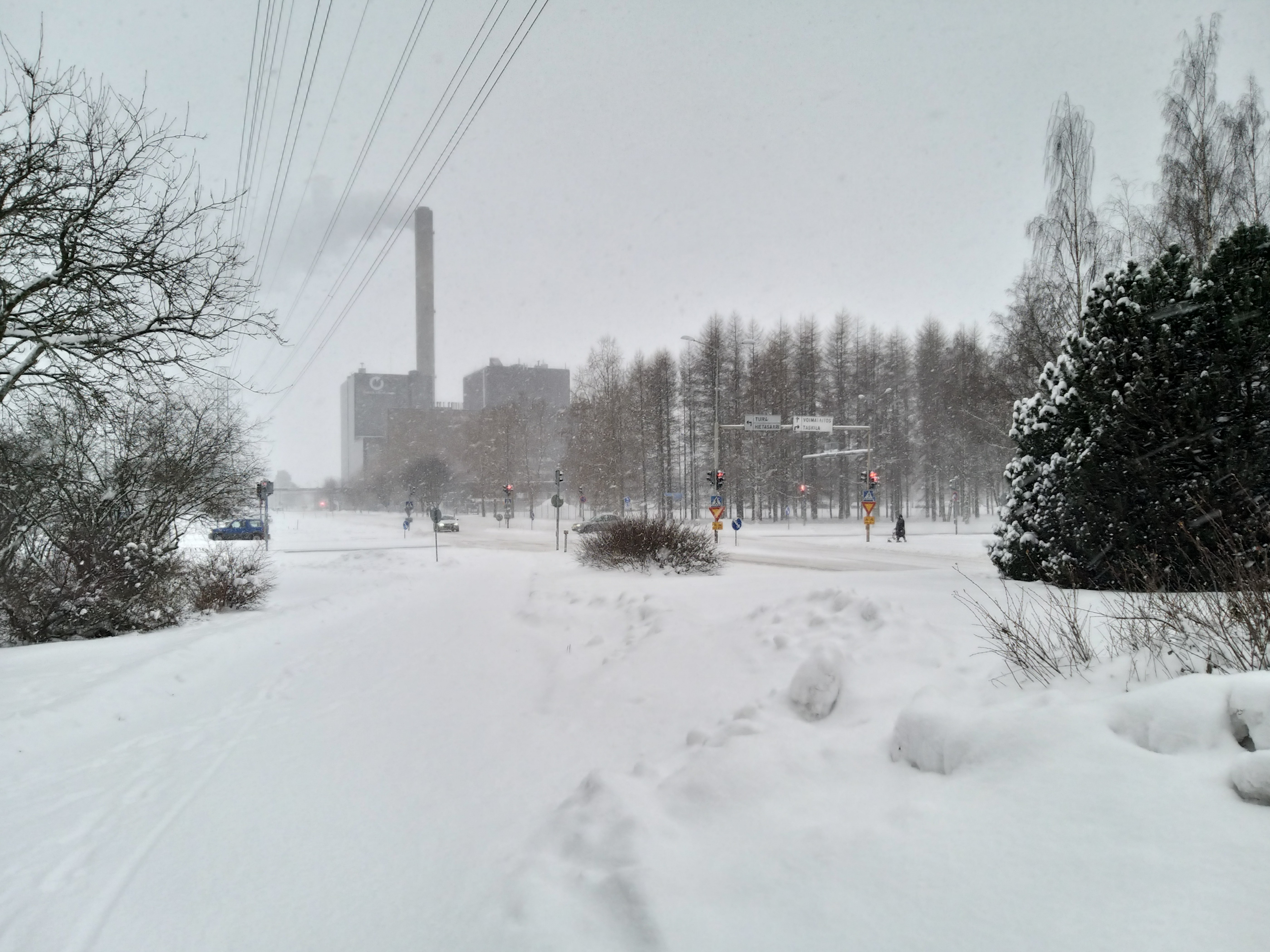 Tervahovintien ja Koskelantien risteys talvella, sakeassa lumisateessa. Taustalla Toppilan voimala. Risteyksessä muutamia autoja sekä yksi jalankulkija rollaattorin kanssa.