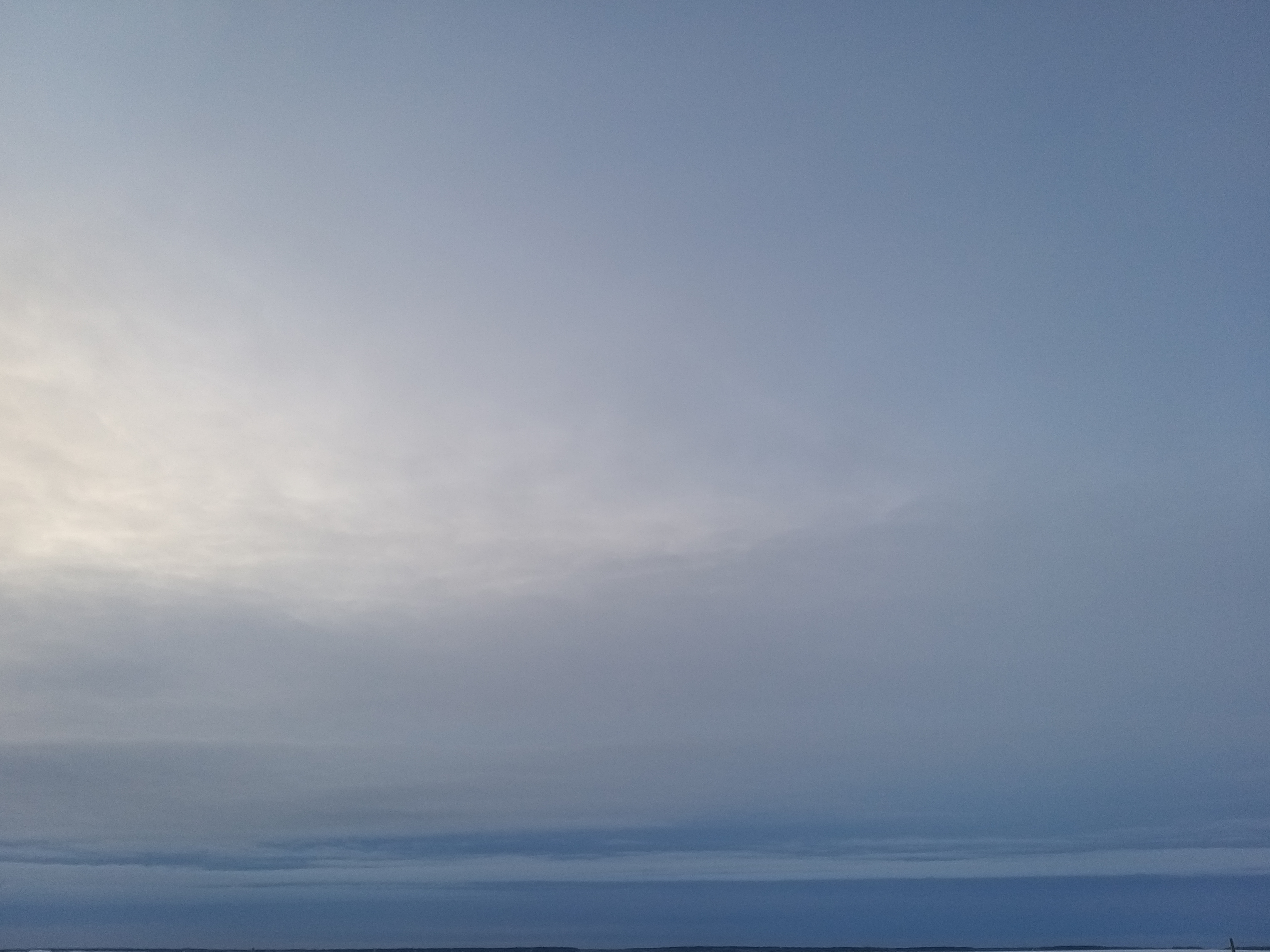 Enimmäkseen pilvinen taivas, vain hieman auringonvalon kajastusta. Ylempänä pilvet harmaita, alhaalla (taivaanrannassa) tummansinisiä.