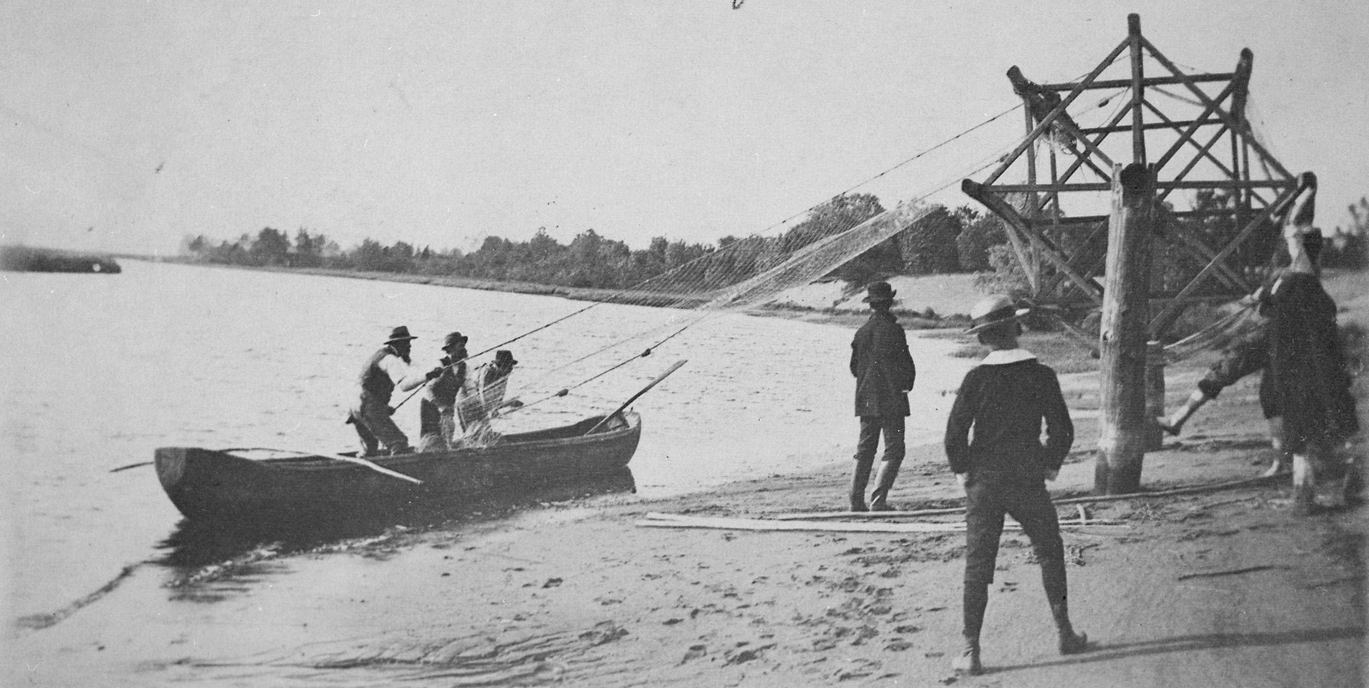 Vanha, mustavalkoinen valokuva: kalaverkkoa kelataan veneestä isolle, rannalla kiinteästi seisovalle puolalle kuivatusta varten.