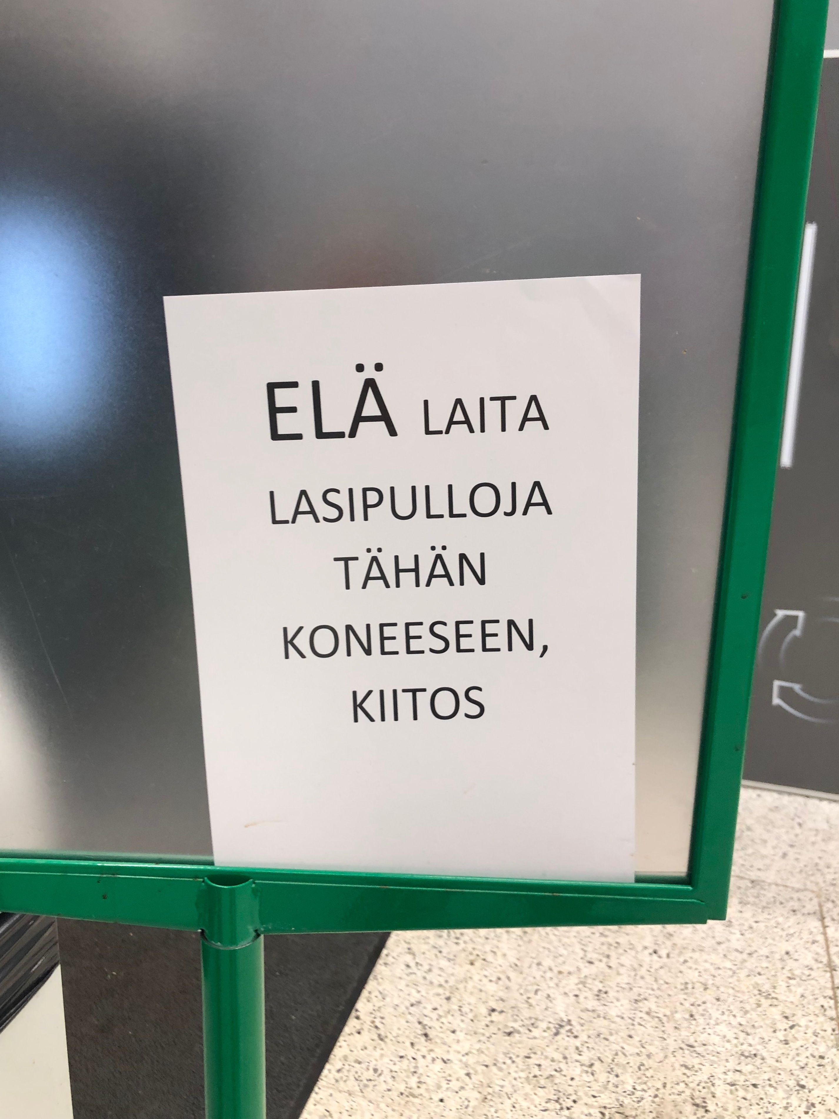Prisman vihreä julisteteline, jonka sisällä tulostettu A4-arkki: "ELÄ laita lasipulloja tähän koneeseen, kiitos"