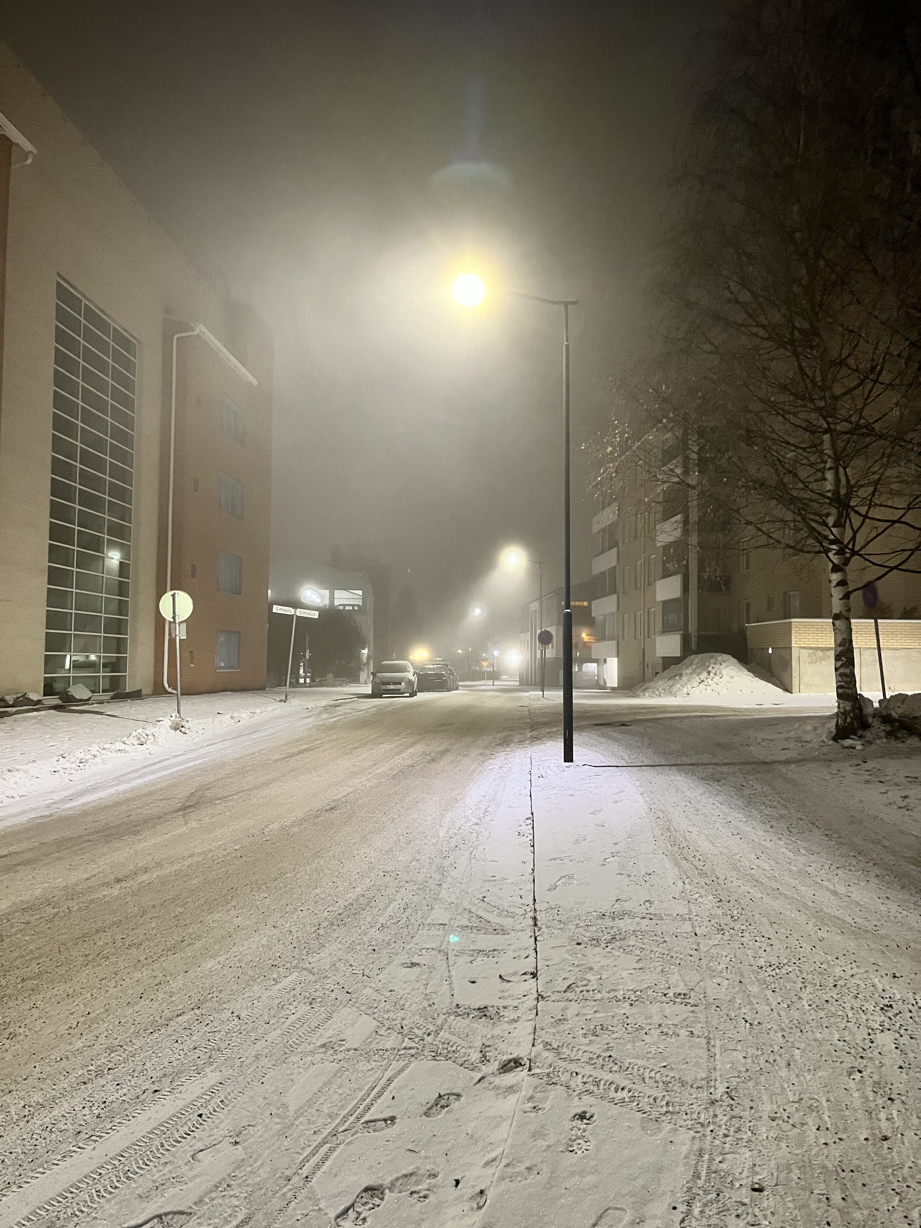 Luminen katu kulkee kerrostalojen välissä. Taivas on pimeä ja katuvalot näkyvät epäterävinä usvan takia.