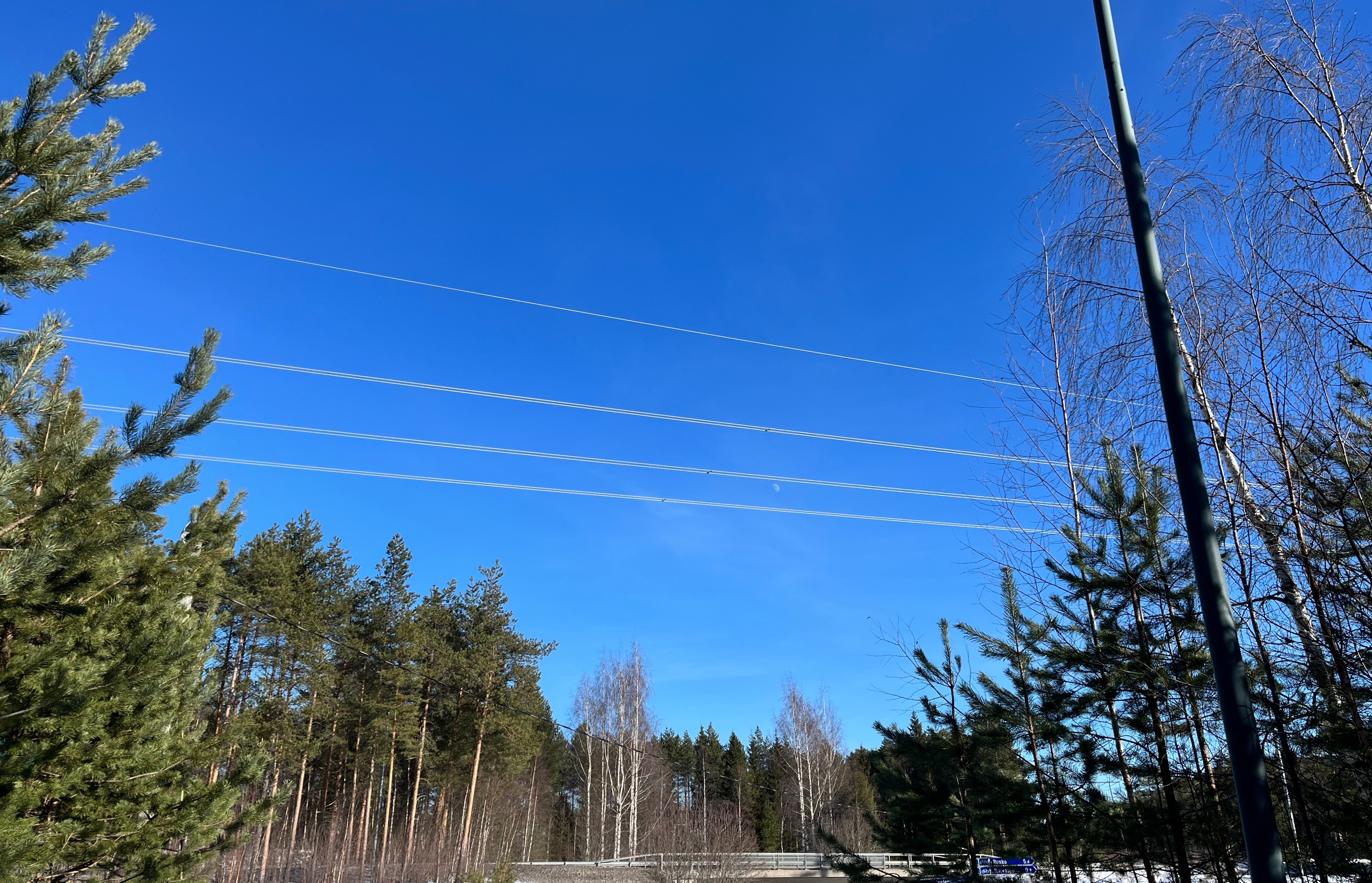 Voimajohtoja kulkee kuvan poikki vasemmalta oikealle. Vasemmalla ja alhaalla (kauempana) puita, enimmäkseen mäntyjä. Aivan alalaidassa (puiden edessä) kuvassa vasemmalta oikealle kulkevan autotien silta. Oikealla valopylväs. Voimajohtojen taustalla on lähes pilvetön, sininen taivas, ja kahden alimman johtonipun välissä puolikuu (kuun oikea puoli näkyy).
