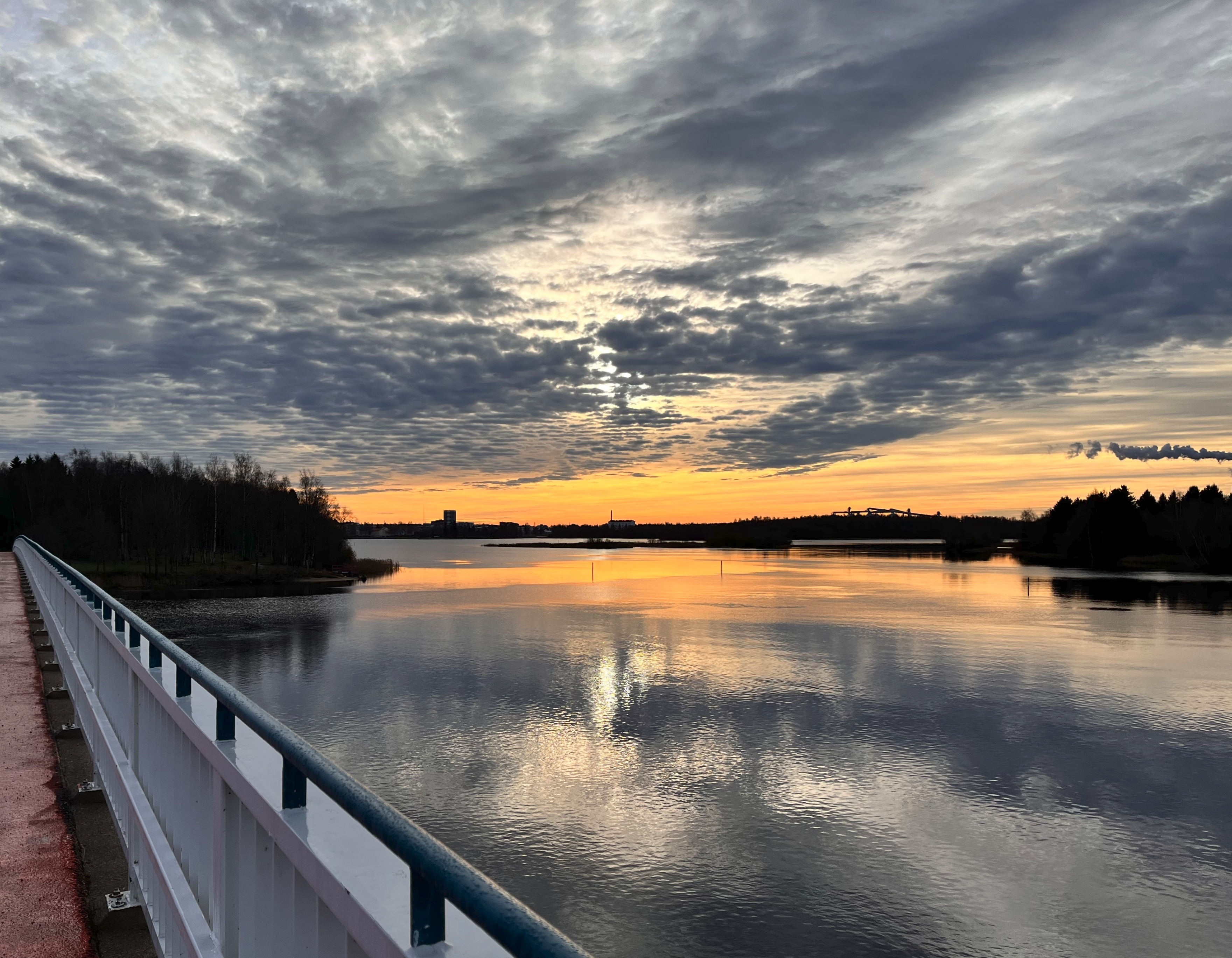 Punertavanoranssi taivaanranta Korkeasaarensillalta kuvattuna. Merenpinta väreilee kevyesti. Aurinko pilkistää siniharmaiden pilvenryppylöiden lomasta.