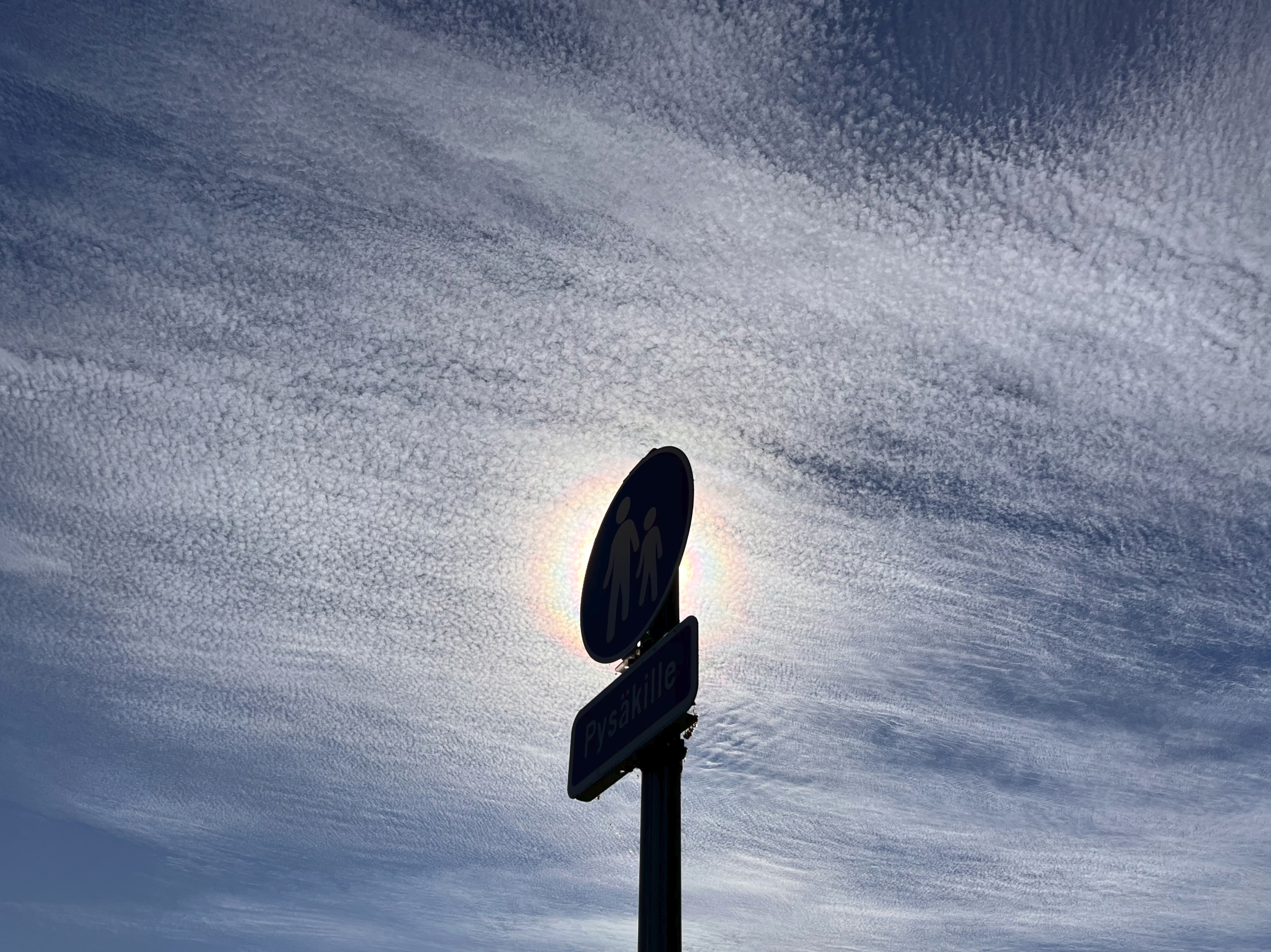 Valkeita palleropilviä (Cirrocumulus) sinisellä taivaalla. Alhaalta kuvan keskelle kohoaa pysäkille vievän jalankulkuväylän liikennemerkki, jonka ylin, pyöreä osa peittää taakseen auringon, niin että auringon värikehä näyttää ympäröivän pyöreää kilpeä. Kilven takana lämpimämpi ilma väreilee niin että siellä palleropilvet näyttävät suttuisemmilta.