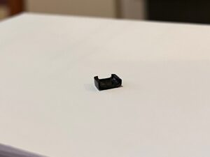 Musta muovinkappale valkealla paperilla kirjainpuoli alaspäin. Sivulta katsottuna muoto on toiselta pitemmältä laidaltaan avoimen laatikon tai kulmikkaan C-kirjaimen kaltainen.