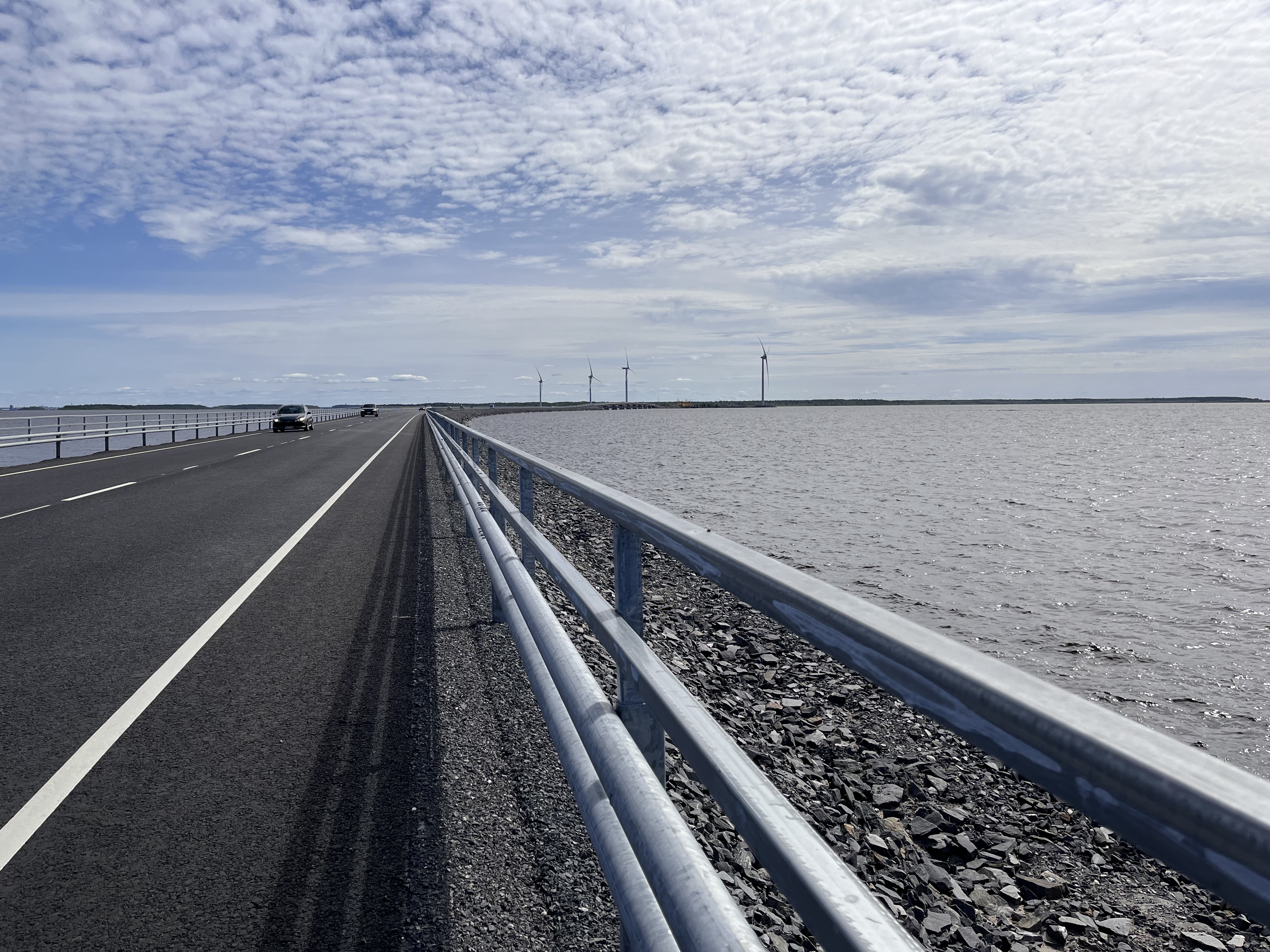 Näkymä Hailuodon siltoja yhdistävän pengertien pientareelta, etelän puolelta, kohti mannerta. Tiellä pari vastaantulevaa autoa. Taivaalla palleropilveä.
